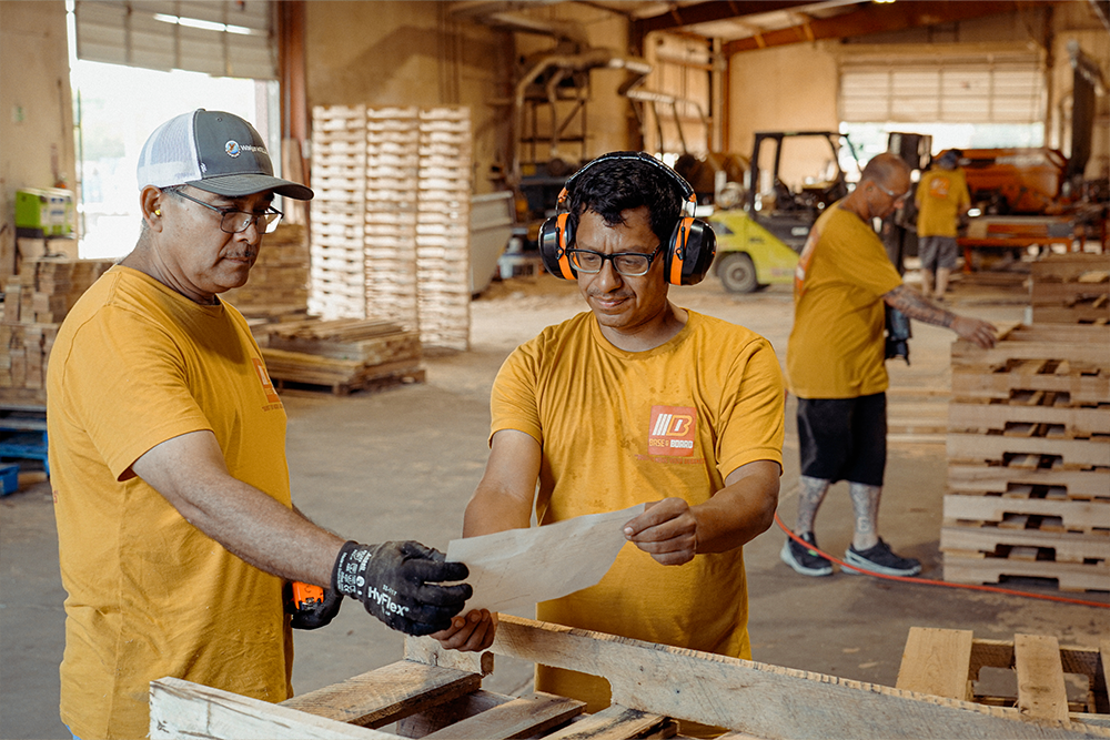 Two workers inspecting documentation in a warehouse with stacked pallets.
